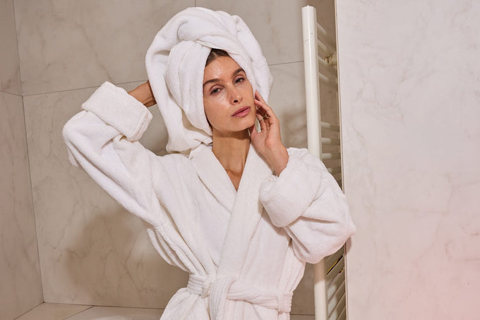 Woman with a towel wrap in a bathroom beside the glowing LumaLux Face V2 LED red light mask